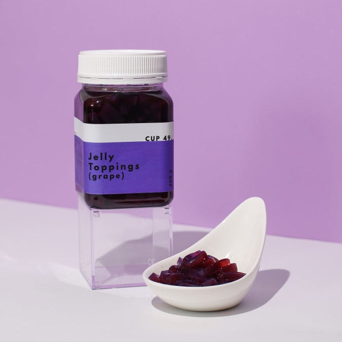 A clear plastic jar containing grape jelly toppings, with a white spoon in front displaying the jelly. The background is purple.
