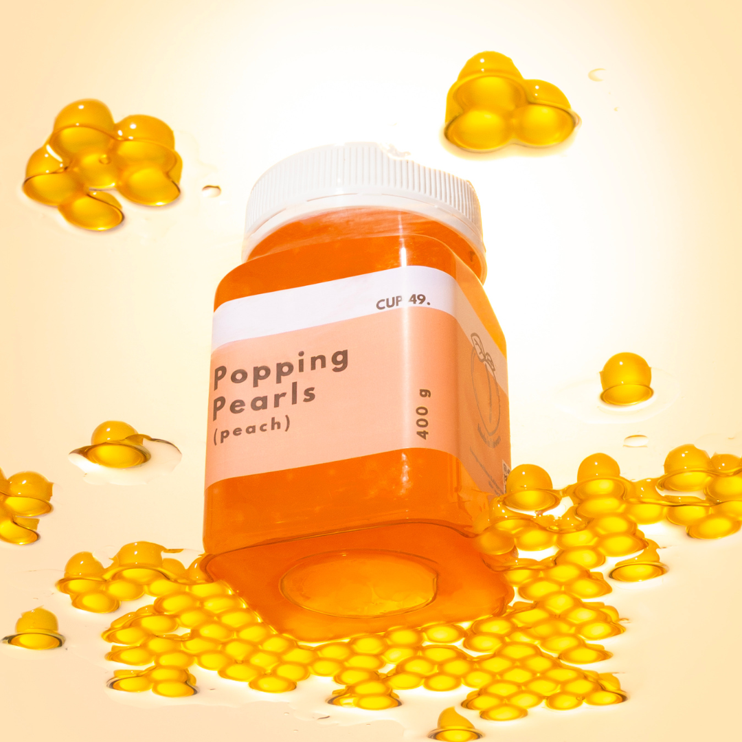 A jar of Popping Pearls peach-flavored popping candy is shown on a warm cream background, surrounded by scattered yellow pearls and glossy drops of the candy. The label reads 'Popping Pearls (peach)' and 'CUP 49.